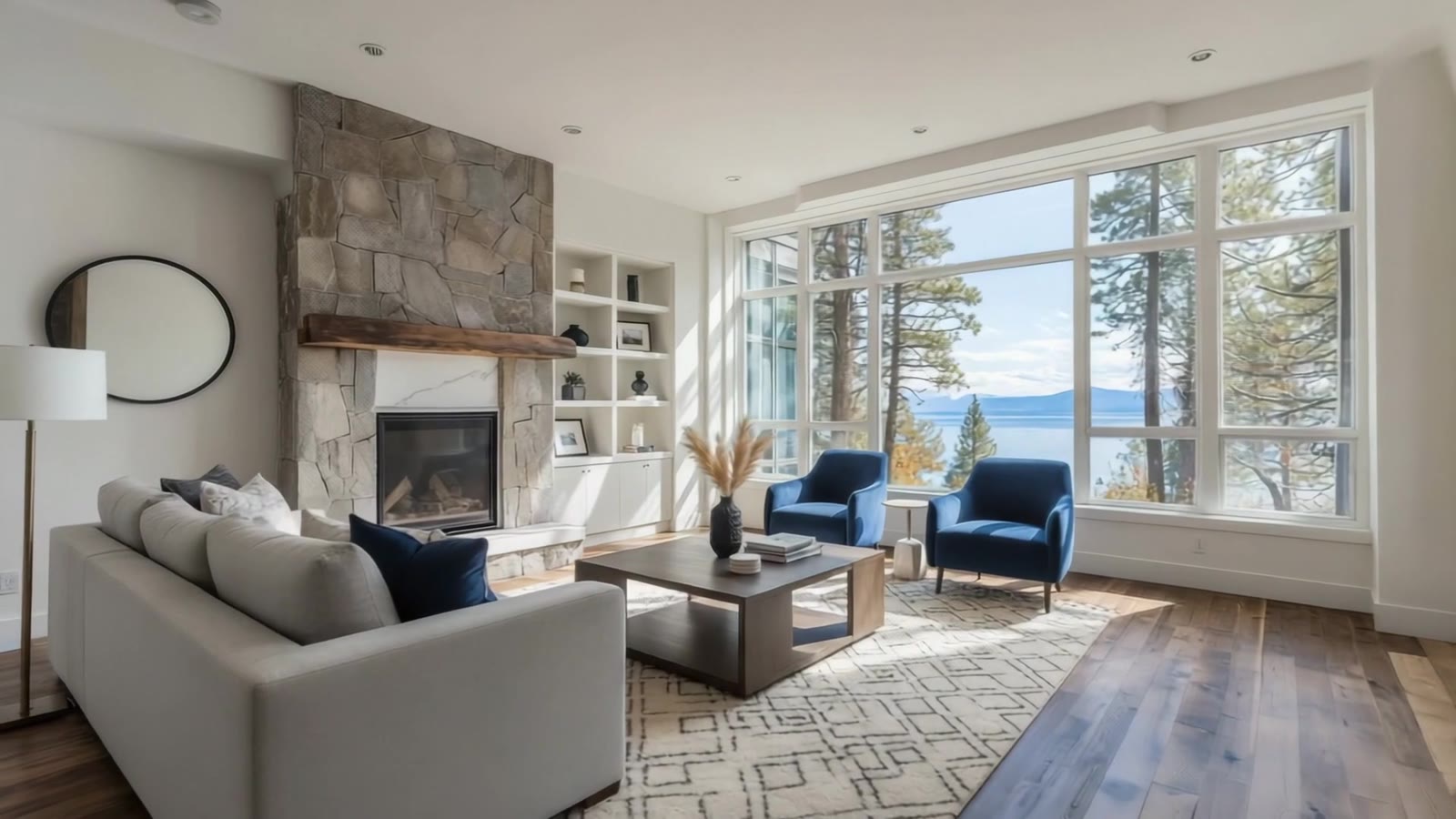 Bright living room with a stone fireplace, blue armchairs and tall windows looking out on pine trees and a lake
